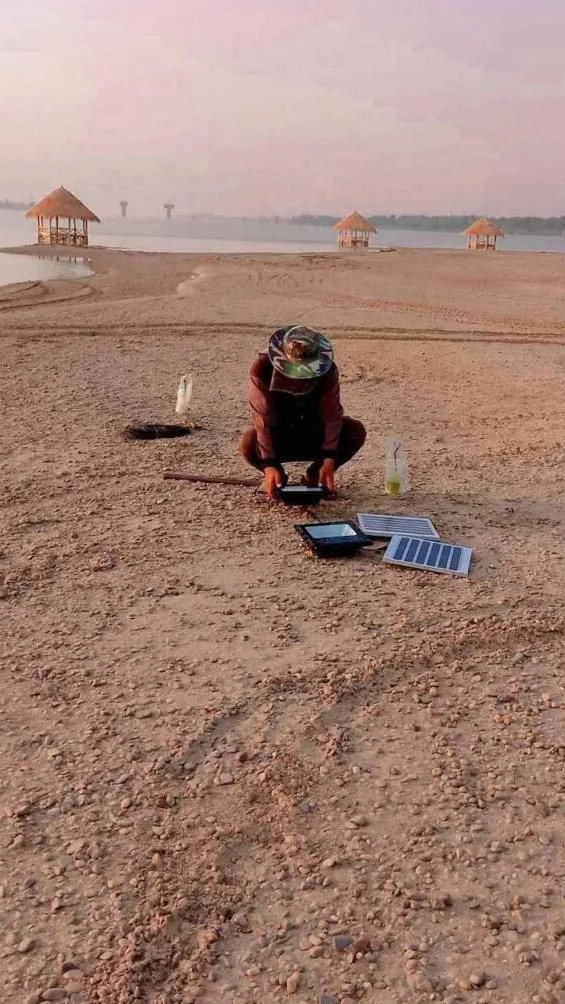 Landscaper crouching on sand fixing a broken solar panel light near the Mekong river.
