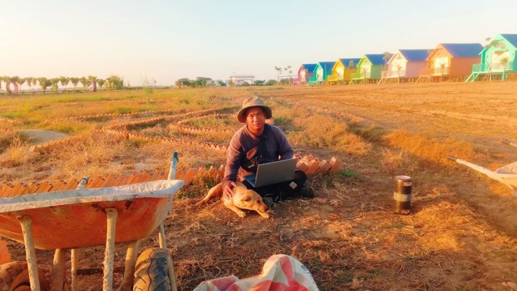 Entrepreneur working on a laptop in the dirt next to a rusty wheelbarrow and dog during resort construction.