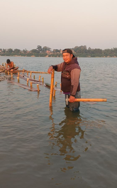 Founder Phonh standing knee-deep in the Mekong River holding a hammer to build a bamboo floating bridge.