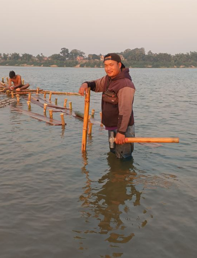 Man standing waist-deep in water holding a large wooden mallet building a bamboo bridge for Dream Garden.