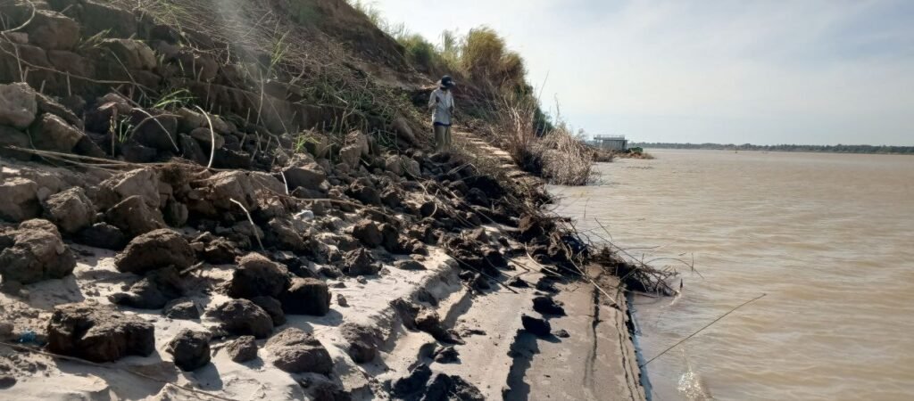 A fisherman standing on a steep, rocky riverbank next to a brown river.