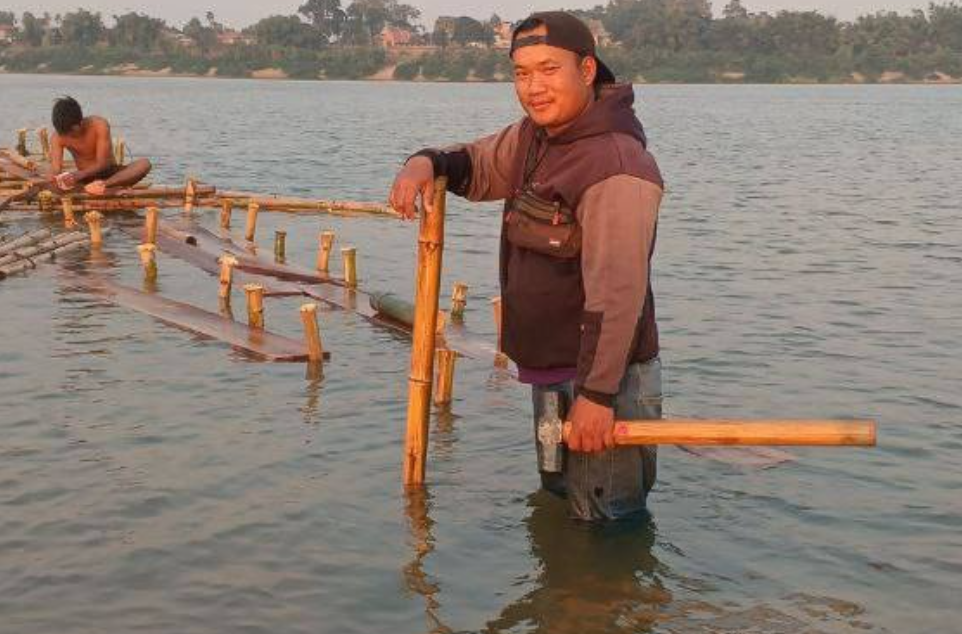 Founder Phonh standing knee-deep in the Mekong River holding a hammer to build a bamboo floating bridge.