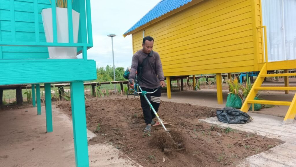 Man using a soil tiller machine to prepare the ground next to a yellow garden cottage.